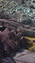 A rocky landscape featuring layers of sedimentary rock with varying textures and colors. Smooth stones and pebbles are scattered across the upper portion, while the foreground displays larger rock formations with visible striations and deep, earthy hues.