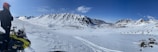 A person wearing a helmet stands beside a snowmobile on a snowy plateau overlooking a vast landscape. Snow-covered mountains rise in the background under a clear blue sky. The untouched snow and the expansive view give a sense of isolation and adventure.