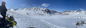 A person wearing a helmet stands beside a snowmobile on a snowy plateau overlooking a vast landscape. Snow-covered mountains rise in the background under a clear blue sky. The untouched snow and the expansive view give a sense of isolation and adventure.
