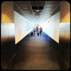 A long corridor with three people walking in the distance, each pulling suitcases. The corridor has a modern design with a dark carpeted floor and white walls. Overhead lights line the ceiling, and narrow windows on the left side bring in natural light.