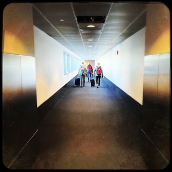 A long corridor with three people walking in the distance, each pulling suitcases. The corridor has a modern design with a dark carpeted floor and white walls. Overhead lights line the ceiling, and narrow windows on the left side bring in natural light.
