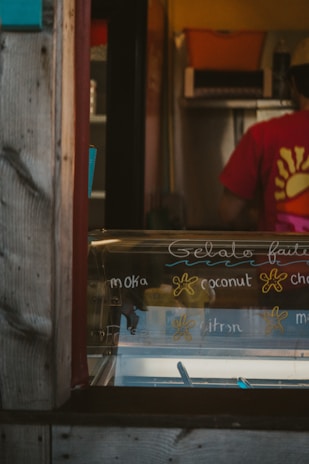 An ice cream shop interior featuring a display case with handwritten gelato flavors like moka, coconut, and citron. A person wearing a red shirt with a yellow sun design is partially visible in the background, standing behind the counter. The shop has a rustic appearance with wooden frame elements.