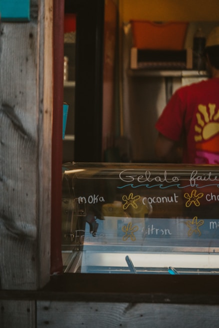An ice cream shop interior featuring a display case with handwritten gelato flavors like moka, coconut, and citron. A person wearing a red shirt with a yellow sun design is partially visible in the background, standing behind the counter. The shop has a rustic appearance with wooden frame elements.