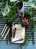 A composition of gardening tools, a book with colorful lettering, a pen, and various plants arranged on a wooden surface. The tools include a small pruning shear and three mini rake tools. The book cover reads 'Wild and Precious Life' in colorful, decorative text. Surrounding the items are green potted plants, including a succulent and a flowering plant with bright pink blossoms.