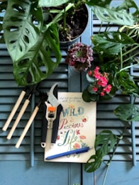 A composition of gardening tools, a book with colorful lettering, a pen, and various plants arranged on a wooden surface. The tools include a small pruning shear and three mini rake tools. The book cover reads 'Wild and Precious Life' in colorful, decorative text. Surrounding the items are green potted plants, including a succulent and a flowering plant with bright pink blossoms.
