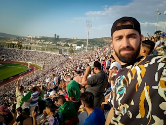 A large crowd fills a stadium, creating a lively and energetic atmosphere. In the foreground, a man takes a selfie, while in the background, the stadium seats are packed with spectators who are watching an event on the field.