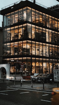 A modern multi-story building with large glass windows, illuminated warmly from the inside. Numerous floors are visible, showcasing a luxurious interior with wooden accents and book-filled shelves. The building is situated on a city street with wet roads, suggesting recent rain. Cars are parked outside, and a small archway marked by lights is visible.