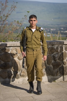 A person in a green military uniform stands confidently on a stone patio with a lush green landscape in the background. The individual holds a rifle in their right hand. The setting is outdoors with clear weather, allowing for a wide view of the scenery.