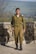 A person in a green military uniform stands confidently on a stone patio with a lush green landscape in the background. The individual holds a rifle in their right hand. The setting is outdoors with clear weather, allowing for a wide view of the scenery.