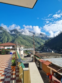 A spacious terrace overlooks a scenic view of lush, green mountains partially covered with clouds under a bright blue sky. The terrace is adorned with potted plants and has a distinctive checkered tile pattern. Various household items, including drying clothes, are visible on the terrace. The surrounding area includes rooftops and utility poles, with a small winding road leading into the town nestled in the hills.