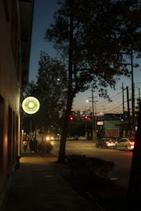 A dimly lit street scene during dusk with a brightly illuminated dental shop sign hanging on the side of a building. Several cars are stopped at a red traffic light, while some pedestrians walk along the sidewalk. Tall trees partially obscure the view of the sky, and various power lines crisscross above the road.