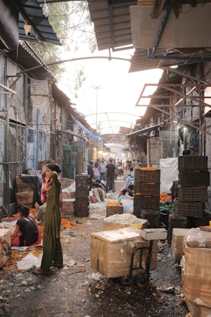 A bustling outdoor market scene with narrow pathways lined by vendors, some seated on the ground surrounded by baskets and trays of vibrant flowers. People walk through the market, with dim evening light filtering through the partially covered roof. Stacks of crates and containers are visible along the sides of the pathways.