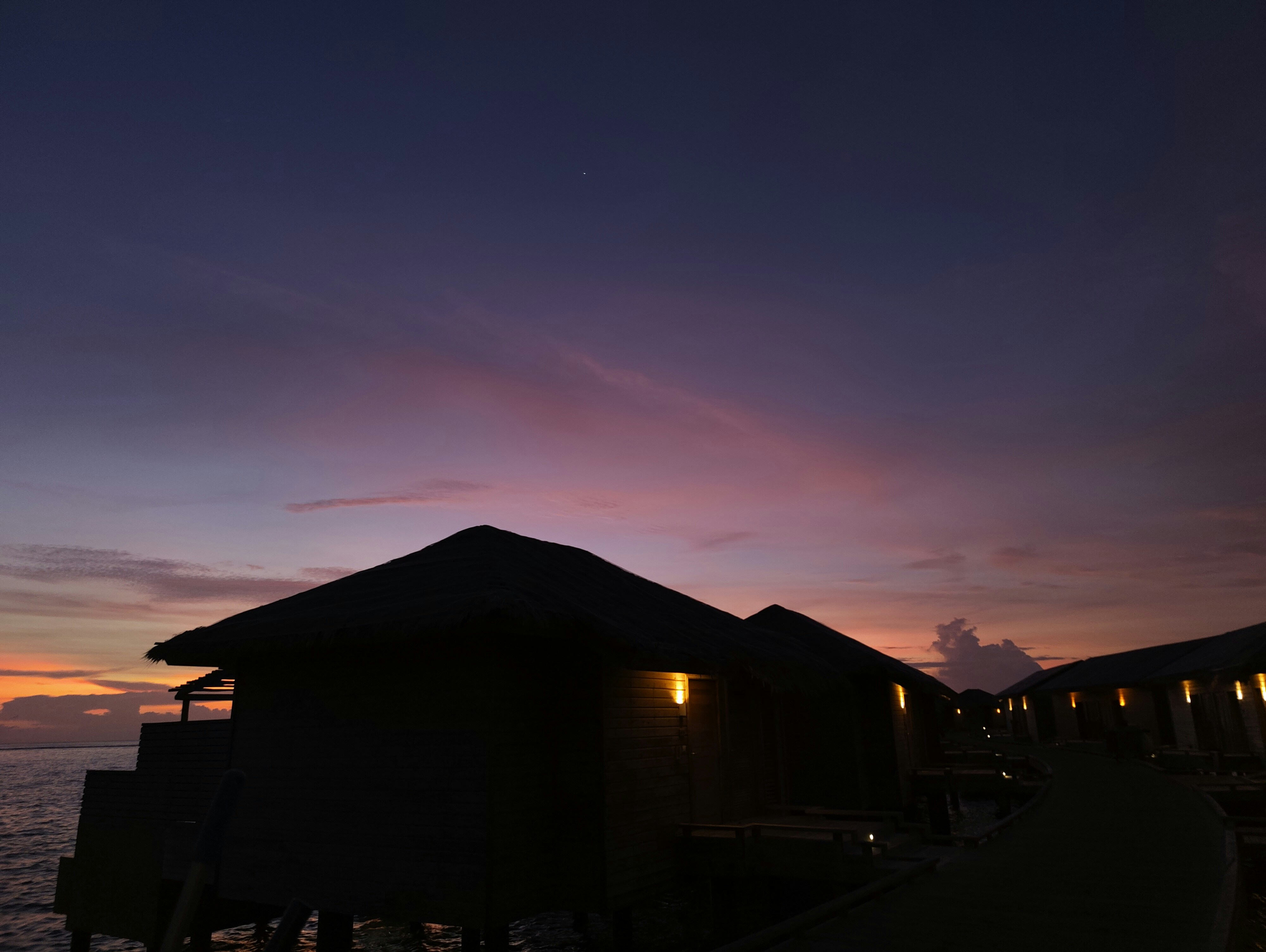 Overwater bungalow at night with dark ocean surrounding, moody deep blue water, dim lantern glow, atmospheric tropical darkness