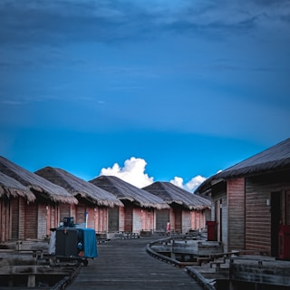 A series of overwater wooden bungalows with thatched roofs aligned along a wooden walkway extending into the distance, set against a backdrop of a vibrant blue sky and fluffy white clouds.