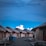 A series of overwater wooden bungalows with thatched roofs aligned along a wooden walkway extending into the distance, set against a backdrop of a vibrant blue sky and fluffy white clouds.