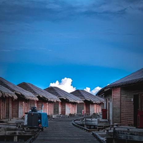 A series of overwater wooden bungalows with thatched roofs aligned along a wooden walkway extending into the distance, set against a backdrop of a vibrant blue sky and fluffy white clouds.