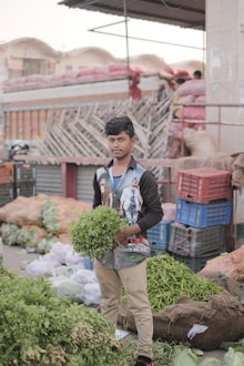 A young man is holding a bunch of fresh greenery, likely coriander, in what appears to be a bustling market setting. In the background, there are piles of vegetables, some packed in bags and crates. The area is filled with assorted produce including green chilies and other leafy vegetables, suggesting a busy, lively market atmosphere.