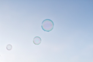 Several transparent bubbles float gracefully against a clear, light blue sky. The bubbles are iridescent, displaying a subtle spectrum of colors around their edges as they catch the light.