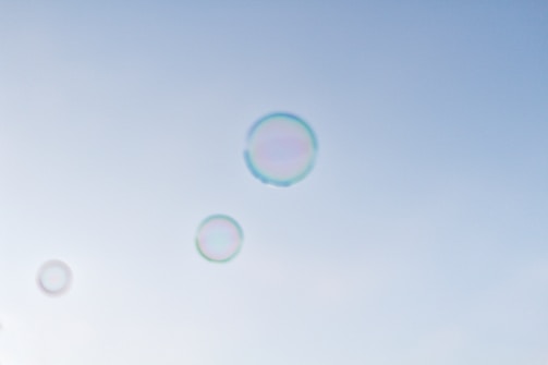 Several transparent bubbles float gracefully against a clear, light blue sky. The bubbles are iridescent, displaying a subtle spectrum of colors around their edges as they catch the light.