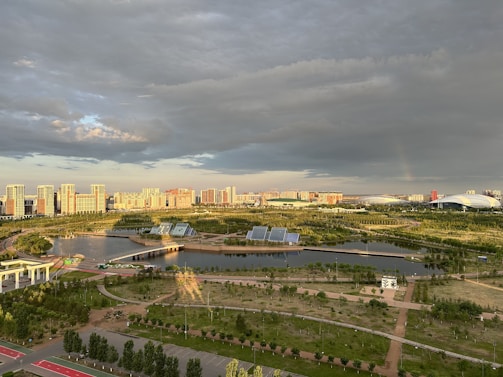 A vibrant cityscape with modern high-rise buildings in the distance, surrounded by lush greenery and a network of roads and pathways. A large body of water with solar panels on platforms is visible in the foreground, along with a variety of trees and grasslands. The sky is a mix of dramatic clouds and sunlight, casting intricate shadows and bright highlights across the landscape.