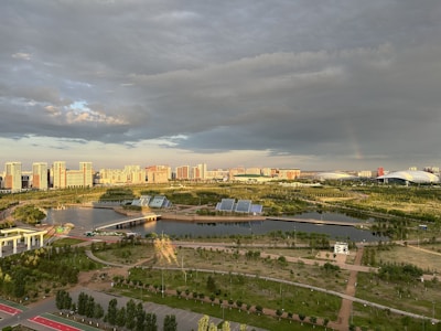 A vibrant cityscape with modern high-rise buildings in the distance, surrounded by lush greenery and a network of roads and pathways. A large body of water with solar panels on platforms is visible in the foreground, along with a variety of trees and grasslands. The sky is a mix of dramatic clouds and sunlight, casting intricate shadows and bright highlights across the landscape.