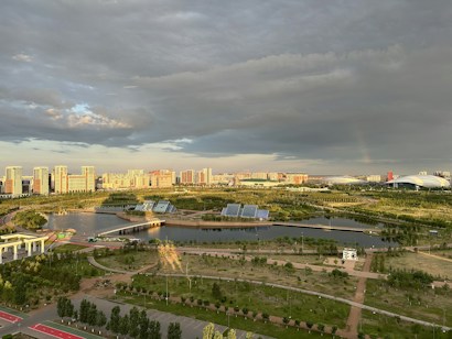A vibrant cityscape with modern high-rise buildings in the distance, surrounded by lush greenery and a network of roads and pathways. A large body of water with solar panels on platforms is visible in the foreground, along with a variety of trees and grasslands. The sky is a mix of dramatic clouds and sunlight, casting intricate shadows and bright highlights across the landscape.