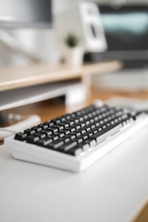 A close-up of a mechanical keyboard with black keys on a white base sits on a light-colored desk. The background is blurred, showing part of a computer monitor and a plant, creating a soft-focus effect.