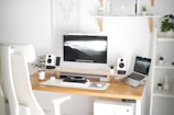 A modern workspace setup with a large monitor displaying a landscape image, a laptop, white speakers, and several small potted plants. The desk is wooden and minimalist, with a white office chair in the foreground. Shelves on the wall hold a few decorative items and more plants, creating a clean and organized environment.