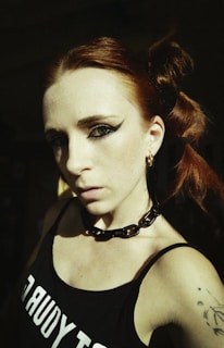 A person with red hair styled in elaborate buns is wearing bold makeup, including thick eyeliner. They are dressed in a black tank top with visible white lettering and a chunky black chain necklace. The lighting casts dramatic shadows, highlighting the person’s facial features and creating an intense atmosphere.