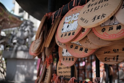 Wooden plaques featuring images of lucky cat figurines and Japanese writing hang from strings, possibly at a temple or shrine setting. A stone statue can be seen blurred in the background, suggesting a sacred or cultural location.