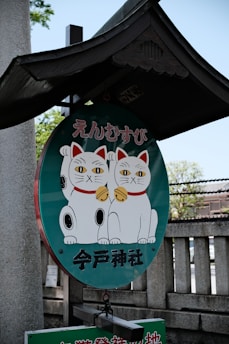 A circular sign features two illustrated white cats with raised paws, wearing red collars and golden bells, which are characteristic of maneki-neko or beckoning cats. The sign is written in Japanese characters. It is attached to a wooden frame resembling a traditional Japanese shrine roof.