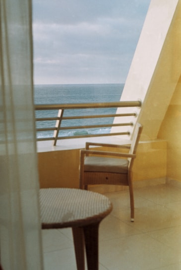 Sunlit balcony with seating area overlooking the ocean from a hotel room.