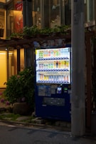 A brightly lit vending machine stocked with various beverages, positioned on a city sidewalk at night. It is surrounded by some greenery, and a tall concrete pillar partially obstructs the view on the right side. The machine features a digital payment option and is filled with colorful drink cans and bottles.