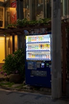 A brightly lit vending machine stocked with various beverages, positioned on a city sidewalk at night. It is surrounded by some greenery, and a tall concrete pillar partially obstructs the view on the right side. The machine features a digital payment option and is filled with colorful drink cans and bottles.