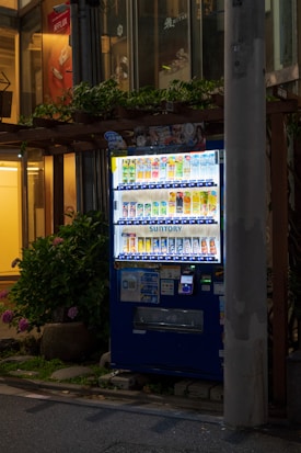 A brightly lit vending machine stocked with various beverages, positioned on a city sidewalk at night. It is surrounded by some greenery, and a tall concrete pillar partially obstructs the view on the right side. The machine features a digital payment option and is filled with colorful drink cans and bottles.