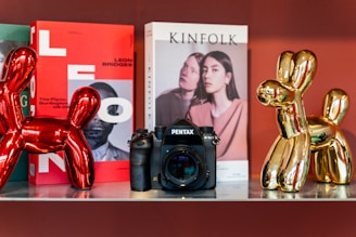 A shelf display featuring a Pentax camera positioned between two metallic balloon dog sculptures, one red and one gold. Behind the camera are two magazines; one has a vibrant red cover with bold letters and an image of a person, while the other has a pale cover with a photograph of two individuals. The scene is set against a warm, reddish background.