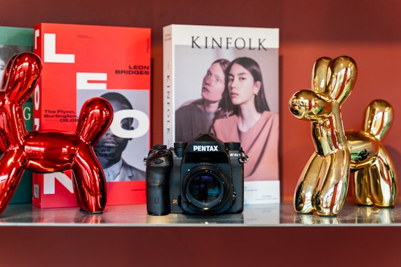 A shelf display featuring a Pentax camera positioned between two metallic balloon dog sculptures, one red and one gold. Behind the camera are two magazines; one has a vibrant red cover with bold letters and an image of a person, while the other has a pale cover with a photograph of two individuals. The scene is set against a warm, reddish background.