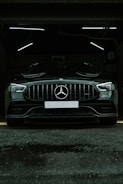 A luxury black car is parked in a dimly lit garage. The sleek vehicle is positioned directly facing the viewer, with its prominent grille and emblem in clear view. The garage's interior lighting creates a stark contrast against the car's glossy finish.