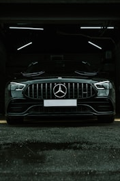 A luxury black car is parked in a dimly lit garage. The sleek vehicle is positioned directly facing the viewer, with its prominent grille and emblem in clear view. The garage's interior lighting creates a stark contrast against the car's glossy finish.