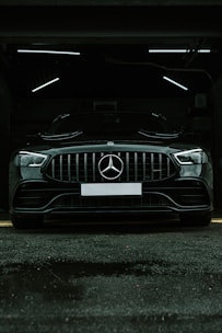 A luxury black car is parked in a dimly lit garage. The sleek vehicle is positioned directly facing the viewer, with its prominent grille and emblem in clear view. The garage's interior lighting creates a stark contrast against the car's glossy finish.