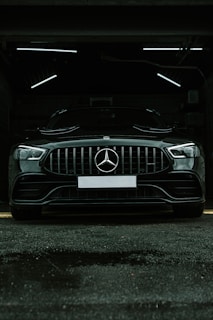 A luxury black car is parked in a dimly lit garage. The sleek vehicle is positioned directly facing the viewer, with its prominent grille and emblem in clear view. The garage's interior lighting creates a stark contrast against the car's glossy finish.