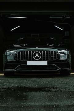 A luxury black car is parked in a dimly lit garage. The sleek vehicle is positioned directly facing the viewer, with its prominent grille and emblem in clear view. The garage's interior lighting creates a stark contrast against the car's glossy finish.