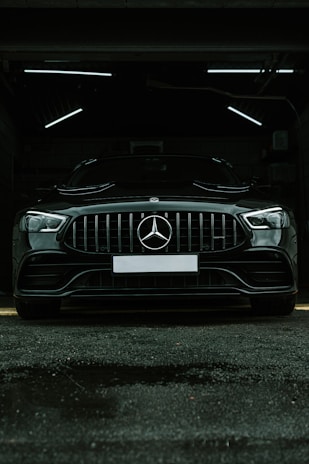 A luxury black car is parked in a dimly lit garage. The sleek vehicle is positioned directly facing the viewer, with its prominent grille and emblem in clear view. The garage's interior lighting creates a stark contrast against the car's glossy finish.