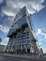 A tall, modern high-rise building under construction with a distinctive geometric design, featuring numerous glass windows reflecting the sky. A large crane is positioned beside the building, and there are construction barriers on the ground level. The sky is partly cloudy, and other buildings can be seen in the distant background.