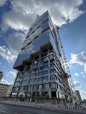 A tall, modern high-rise building under construction with a distinctive geometric design, featuring numerous glass windows reflecting the sky. A large crane is positioned beside the building, and there are construction barriers on the ground level. The sky is partly cloudy, and other buildings can be seen in the distant background.