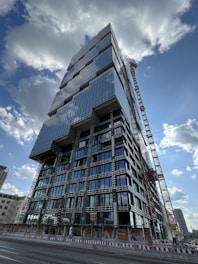 A tall, modern high-rise building under construction with a distinctive geometric design, featuring numerous glass windows reflecting the sky. A large crane is positioned beside the building, and there are construction barriers on the ground level. The sky is partly cloudy, and other buildings can be seen in the distant background.