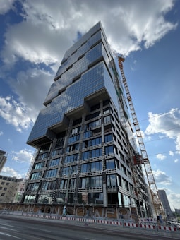 A tall, modern high-rise building under construction with a distinctive geometric design, featuring numerous glass windows reflecting the sky. A large crane is positioned beside the building, and there are construction barriers on the ground level. The sky is partly cloudy, and other buildings can be seen in the distant background.