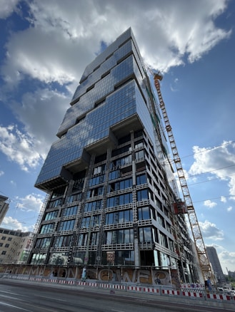 A tall, modern high-rise building under construction with a distinctive geometric design, featuring numerous glass windows reflecting the sky. A large crane is positioned beside the building, and there are construction barriers on the ground level. The sky is partly cloudy, and other buildings can be seen in the distant background.