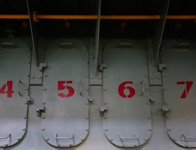A series of metal doors lined up in a row, each numbered sequentially from 4 to 7 with red paint. The metal appears industrial with visible bolts and hinges, and there is a yellow pipe running horizontally above the doors.
