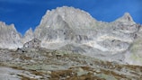 A rustic mountain cabin nestled at the base of towering, rugged granite peaks. The cabin is surrounded by rocky and grassy terrain under a clear blue sky.